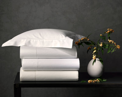 A stack of neatly folded white bed sheets and a pillow sit on a black table beside a small round vase with yellow and white flowers against a gray textured wall.