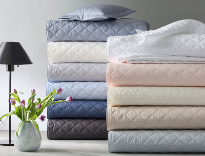 Stacked folded comforters in various pastel and neutral colors, a pillow on top, a black table lamp, and a vase with purple tulips on a gray surface.