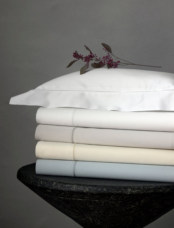 A stack of folded bed sheets in neutral tones topped with a white pillow and a sprig of purple flowers, set against a gray background.