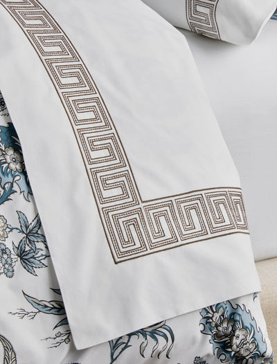 Close-up of a white bedsheet with a brown Greek key border, layered over floral-patterned bedding in blue, black, and white tones.
