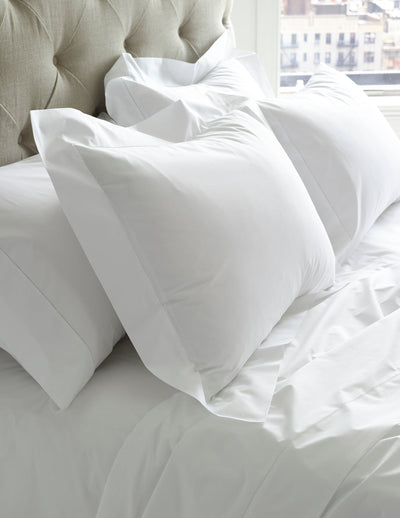 A neatly made bed with white pillows and sheets, set against a tufted headboard and a window showing buildings outside.