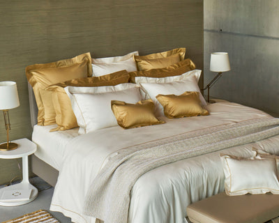 A neatly made bed with white and gold bedding, multiple pillows, a white nightstand with a lamp, and a modern, neutral-colored background.