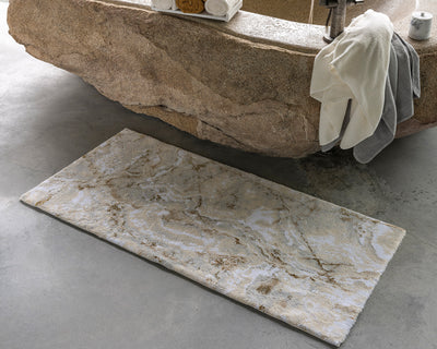 Rectangular bath mat with a marble-like pattern placed on a gray concrete floor in front of a stone bathtub draped with white and gray towels.
