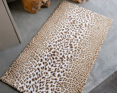 Rectangular rug with a leopard print pattern in brown and white tones placed on a gray concrete floor next to a wooden decor item.
