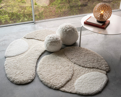 A modern room with two textured, cream-colored rugs, two plush round cushions, and a white table holding books and a lit woven lamp, near a large window overlooking greenery.