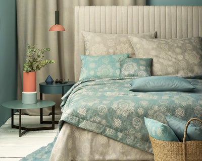 Neatly made bed with blue and beige patterned bedding, flanked by small tables with decorative items, a plant, and a woven basket in a softly lit, modern bedroom.