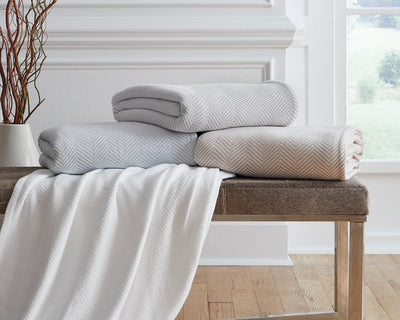 Three folded herringbone-patterned blankets in light gray, beige, and white are stacked on a wooden bench, with one white blanket draped over the side.