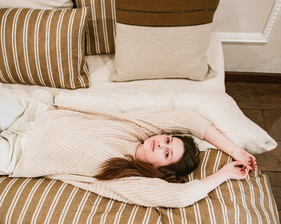 A woman in a cream sweater lies on a bed with striped brown and beige pillows and bedding, looking up with her arms stretched above her head.