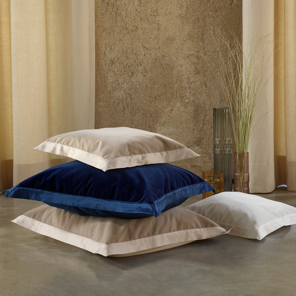 Four large pillows in beige, navy blue, and white stacked on a concrete floor near a glass vase with tall grass, against a textured wall and beige curtains.