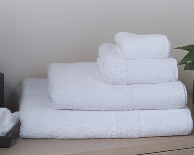 Four neatly folded white towels of varying sizes stacked on a wooden surface with a plant partially visible on the right.
