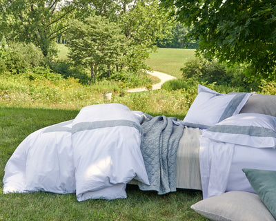 A neatly made bed with white and gray bedding is set outdoors on green grass, with trees and a winding path in the background.