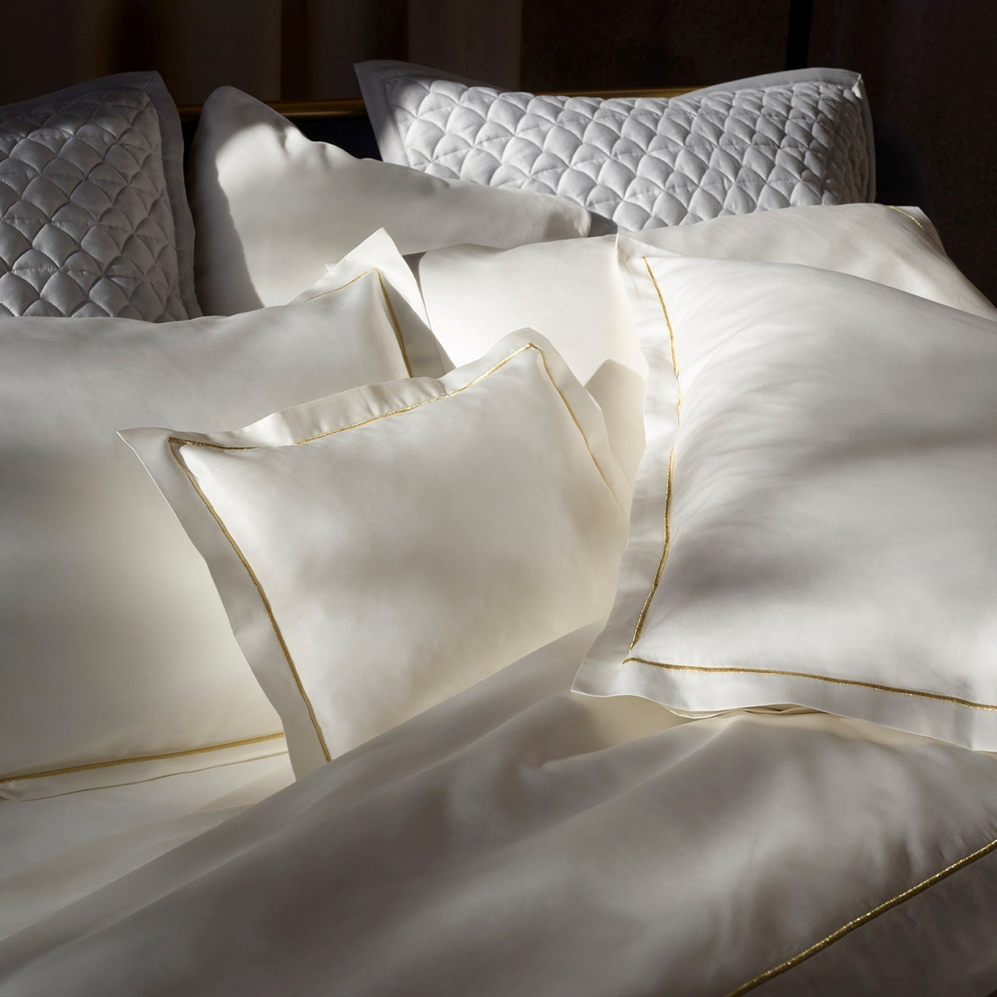 A bed with several white pillows and a white duvet, some with gold piping and some with a quilted pattern, bathed in natural light.