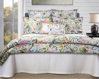 A neatly made bed with a floral-patterned comforter and pillow set, a white textured bedspread, a beige accent pillow, and a lamp on a bedside table.