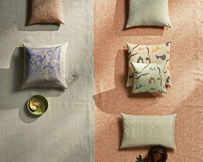 A top view of patterned cushions arranged on two adjacent fabrics, one light gray and one peach, with a green plate and a glass placed on the gray side.