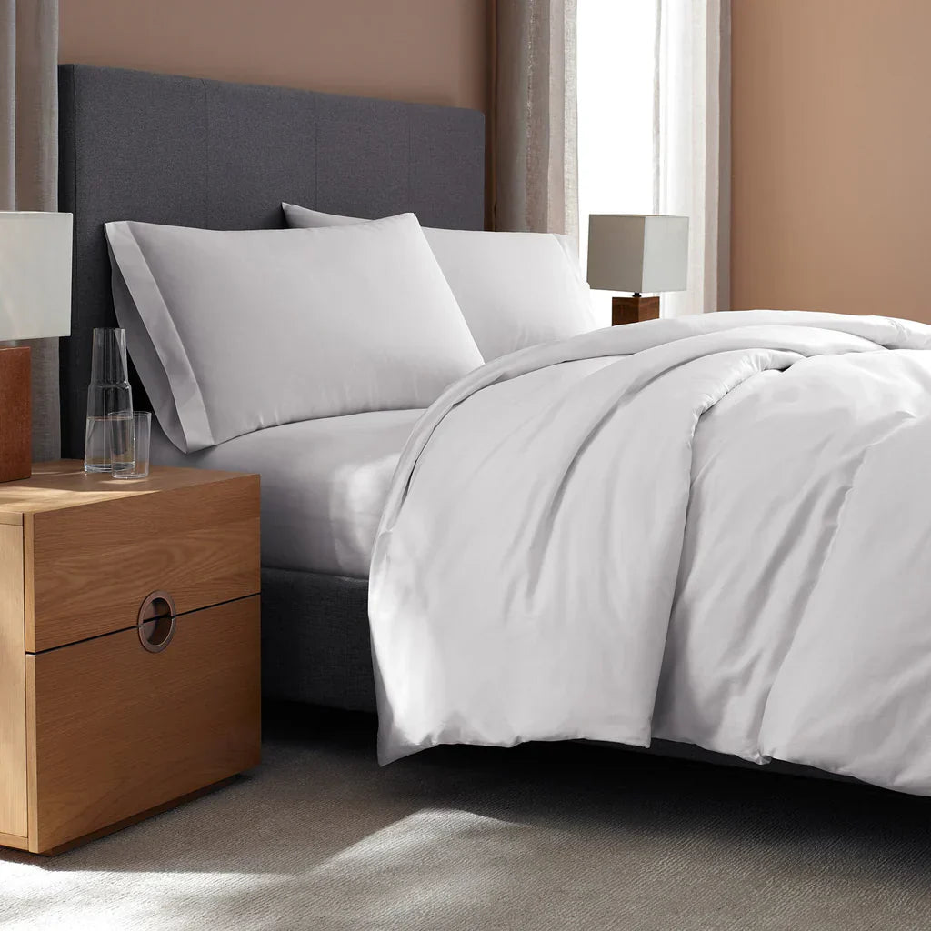 A neatly made bed with white bedding and pillows next to a wooden nightstand with a glass of water and a lamp, in a modern, neutral-toned bedroom.
