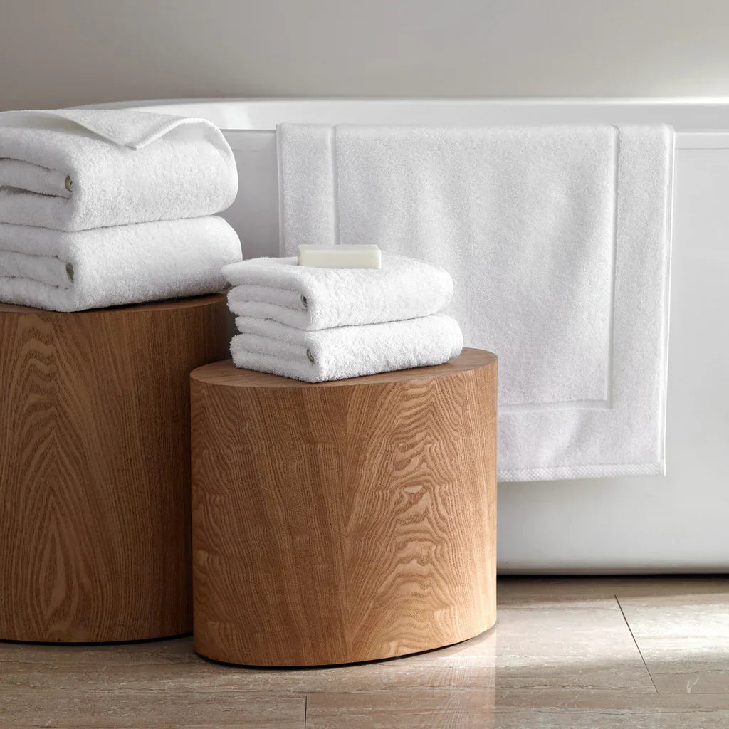 Two stacked wooden stools hold folded white towels and a bar of soap beside a bathtub draped with a white towel on a tiled floor.