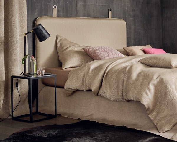 A neatly made bed with beige patterned bedding, several pillows, a black bedside lamp, a small black table, a glass of water, and a flower in a vase.