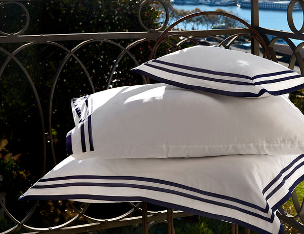 Three white pillows with navy blue trim stacked on an outdoor metal chair, with greenery and water visible in the background.