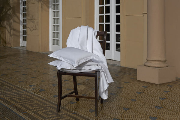 A dark wooden chair on a tiled patio with white bedding and a pillow draped over it, positioned near glass doors and a beige column.
