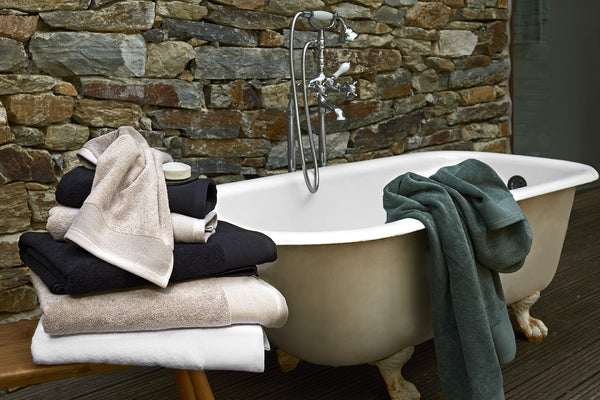 A clawfoot bathtub with a green towel draped over the side, a stone wall background, and a wooden bench stacked with folded towels and a small jar.