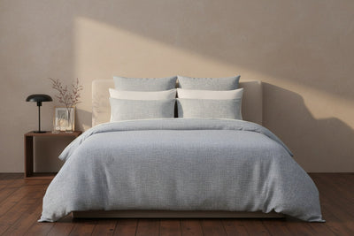 A neatly made bed with light gray bedding is centered in a minimal bedroom, beside a wooden nightstand with a lamp, a vase, and framed art.