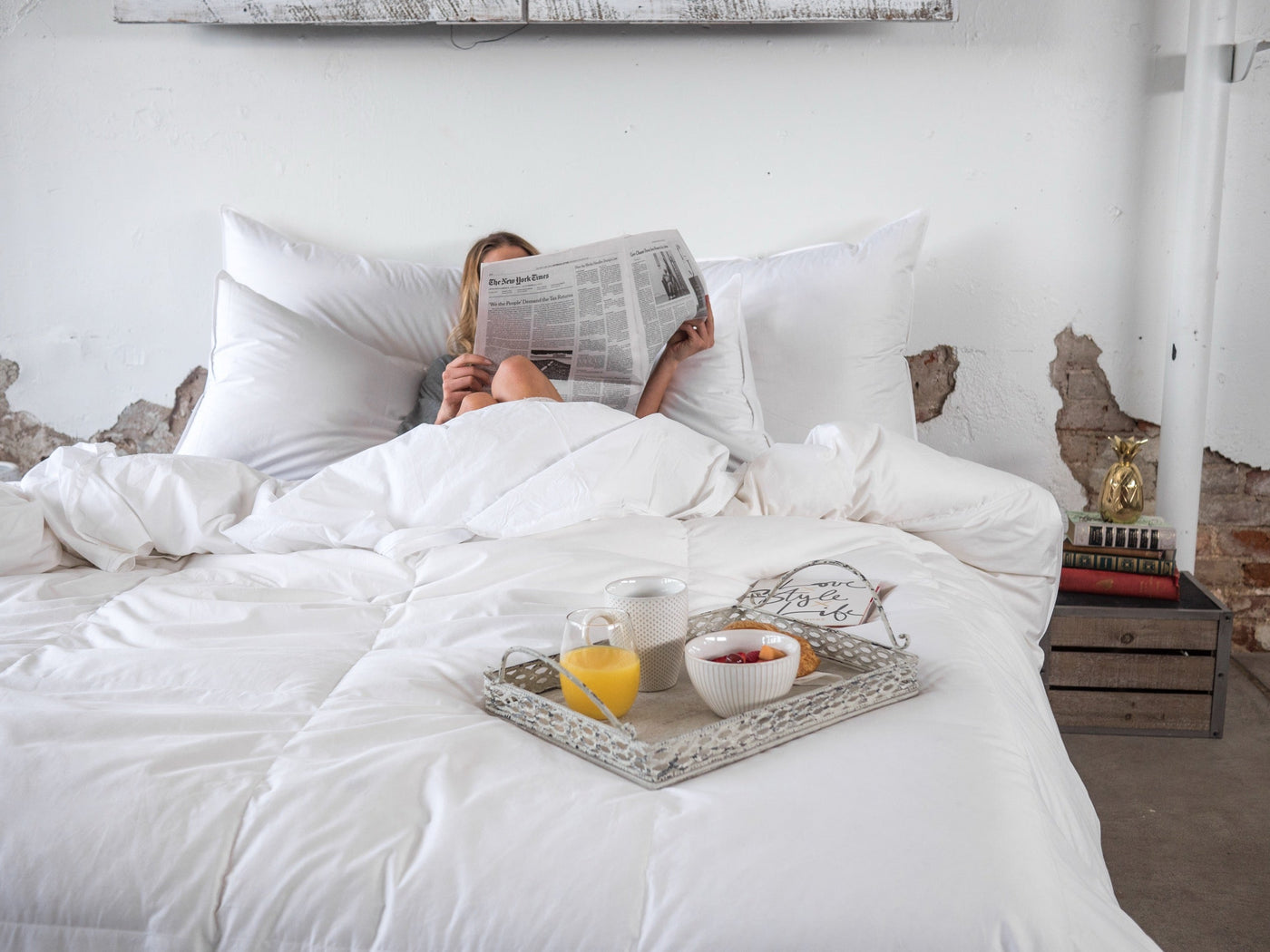 Person reading newspaper in bed with white down duvet, breakfast tray with juice and bowl in foreground.