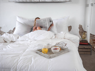 Person reading newspaper in bed with white down duvet, breakfast tray with juice and bowl in foreground.