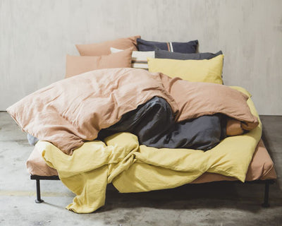 Unmade bed with yellow, peach, and dark gray bedding and several pillows against a plain light-colored wall.
