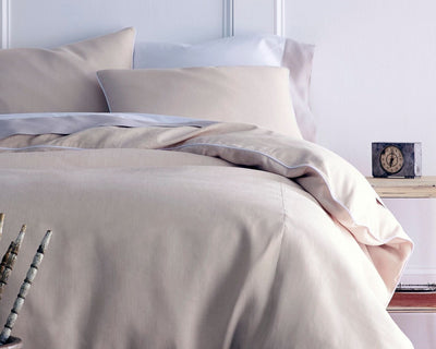 A neatly made bed with beige sheets and pillows next to a small wooden table holding a vintage clock.