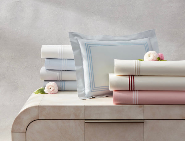 Folded bed sheets and pillowcases in pastel shades, some with stripes, are neatly stacked on a light-colored dresser, with a few flowers placed on top.