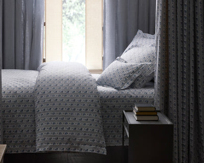 A neatly made bed with patterned blue and white bedding is next to a window with sheer curtains; a small table with stacked books sits beside the bed.
