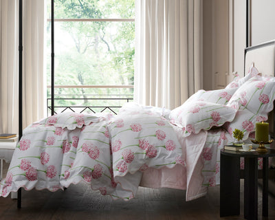 A neatly made bed with white and pink floral bedding sits beside a round black table, in front of a large window with beige curtains and a view of green trees outside.
