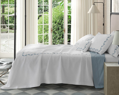 A neatly made bed with white bedding and blue embroidery is positioned beside large windows overlooking a garden; a floor lamp and a side table are nearby.