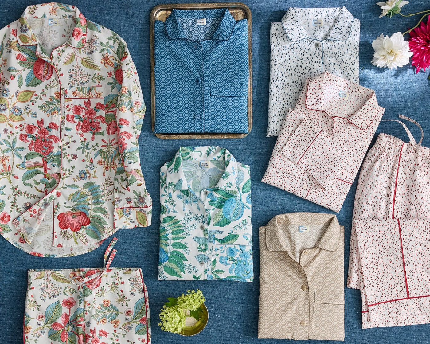 A variety of patterned pajama sets in different colors and floral or geometric designs are arranged neatly on a blue surface, with flowers and a small tray as decoration.