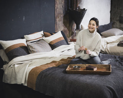 A woman in a light sweater sits on a bed with striped bedding, smiling, with a wooden tray holding a cup and food in front of her in a cozy, dimly lit room.