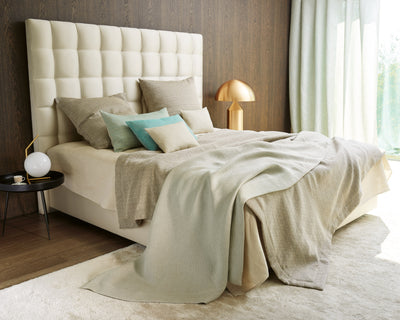 A modern bedroom with a tall, tufted headboard, beige bedding, assorted pillows, a round black side table, and a gold lamp, with natural light from a window.