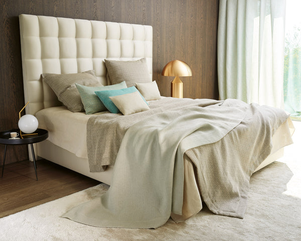 A modern bedroom with a tall, tufted headboard, beige bedding, assorted pillows, a round black side table, and a gold lamp, with natural light from a window.