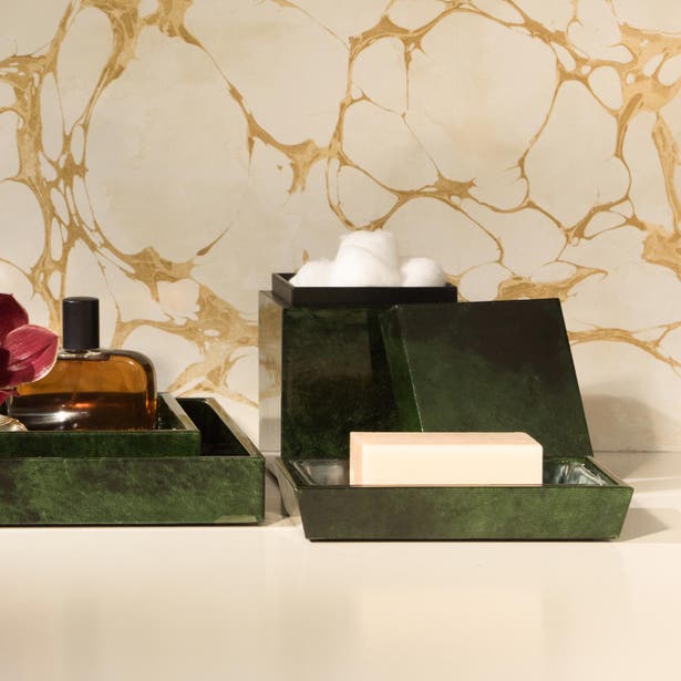 Two dark green lacquered bath boxes styled with soap and cotton balls against a gold marble wall.