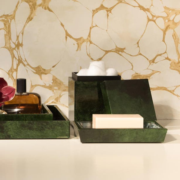 Two dark green lacquered bath boxes styled with soap and cotton balls against a gold marble wall.