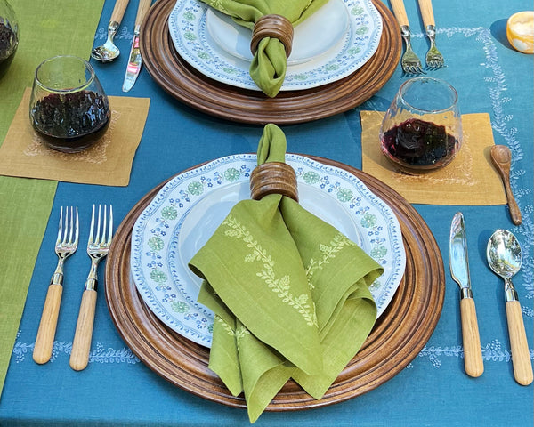 A table setting with wooden chargers, patterned plates, green napkins in wooden rings, wine glasses with red wine, and utensils with wooden handles on a blue tablecloth.