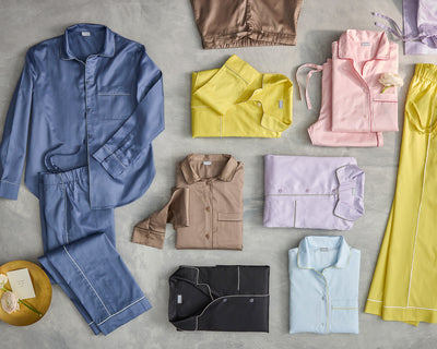 A variety of folded and laid-out pajamas in different colors, including blue, yellow, pink, brown, lavender, black, and light blue, displayed on a gray surface.