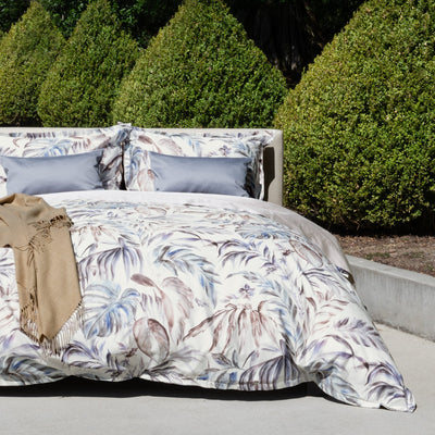 A bed with a floral-patterned duvet and two gray pillows is set outdoors on a concrete patio, with manicured green bushes in the background. A tan blanket is draped on the corner.