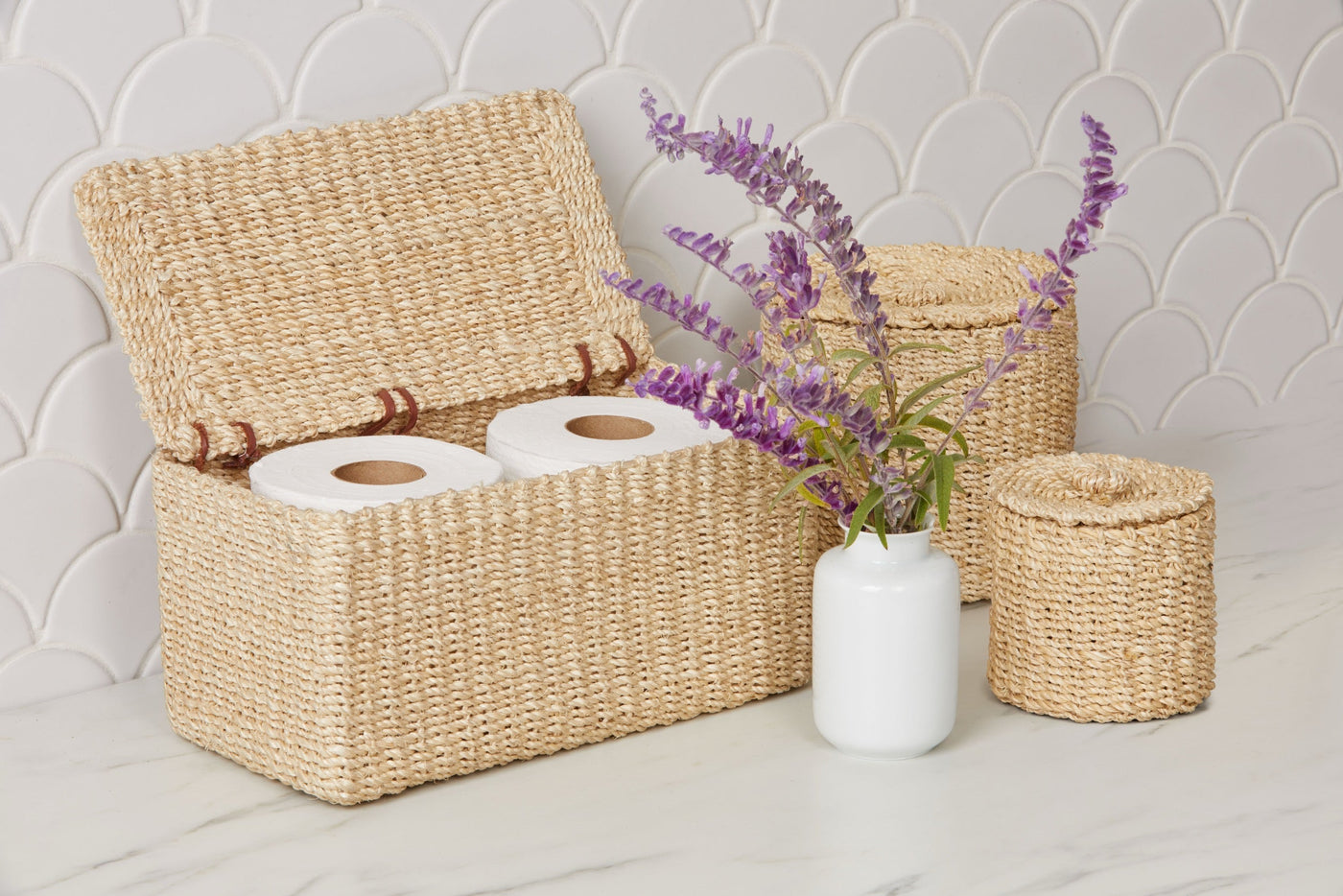 Chelston woven seagrass bath basket set styled with purple florals on marble surface.