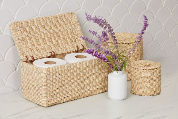 Chelston woven seagrass bath basket set styled with purple florals on marble surface.