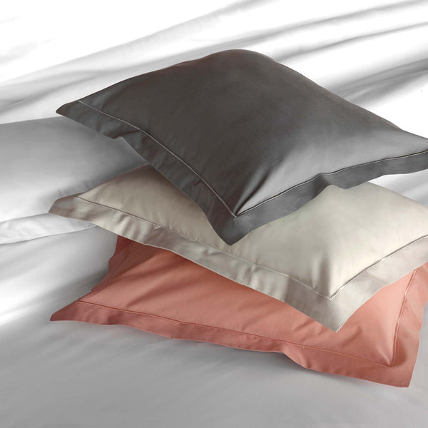 Three neatly stacked pillows in gray, cream, and pink pillowcases rest on a white bed with white sheets and a pillow in the background.