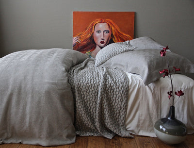 A neatly made bed with gray textured bedding and pillows, a painting of a woman with red hair propped behind it, and a vase with red flowers on the wooden floor.