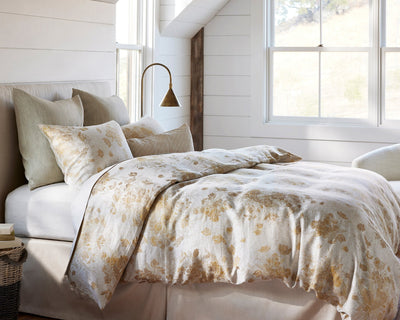 A neatly made bed with light-colored floral bedding, beige and gray pillows, a brass reading lamp, and large windows in a bright, airy bedroom.
