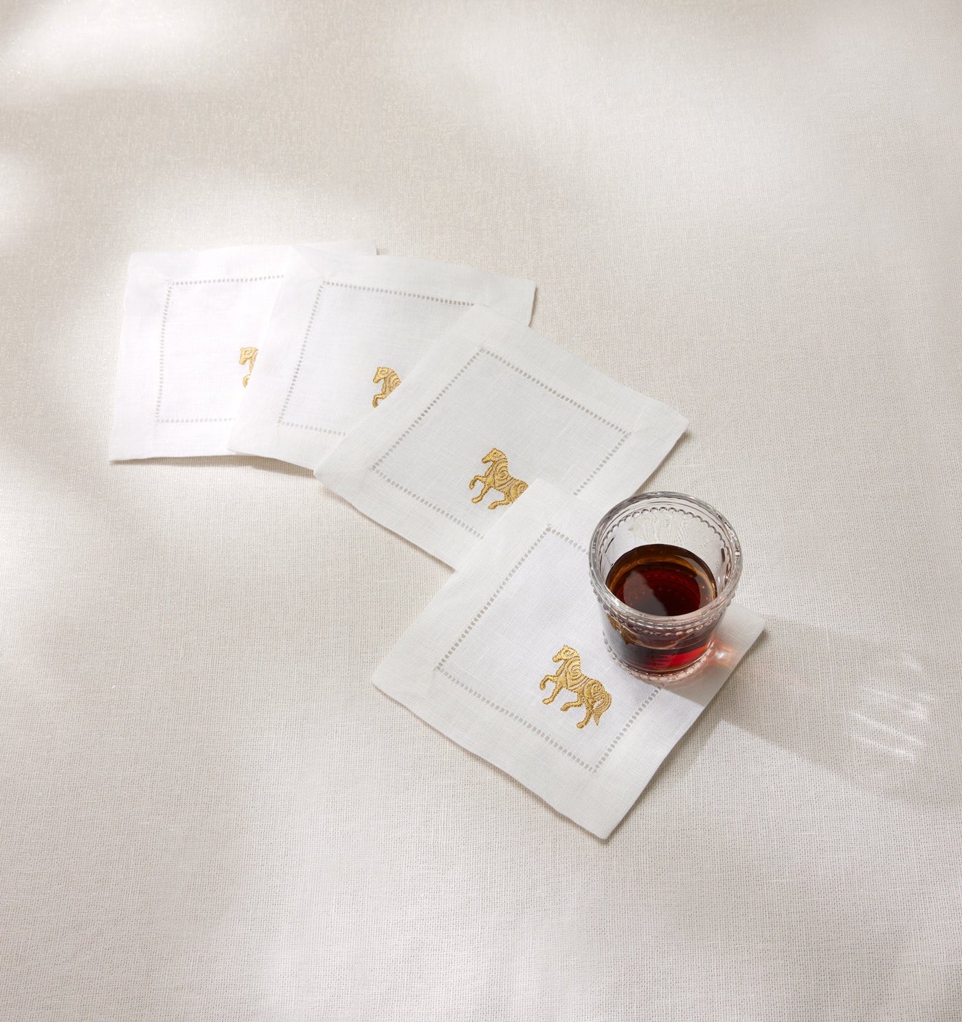 Ivory linen cocktail napkins with gold embroidered horse motif, styled with a glass of red wine.