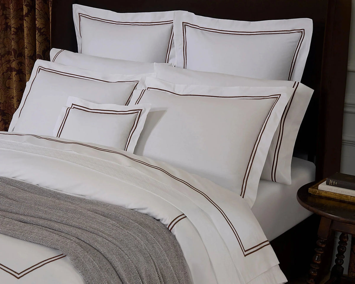White bedding set with brown embroidered border details, styled on a bed with a dark brown headboard, a gray throw blanket, and a bedside table in a bedroom setting.
