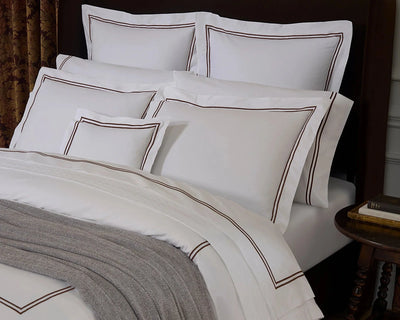 White bedding set with brown embroidered border details, styled on a bed with a dark brown headboard, a gray throw blanket, and a bedside table in a bedroom setting.